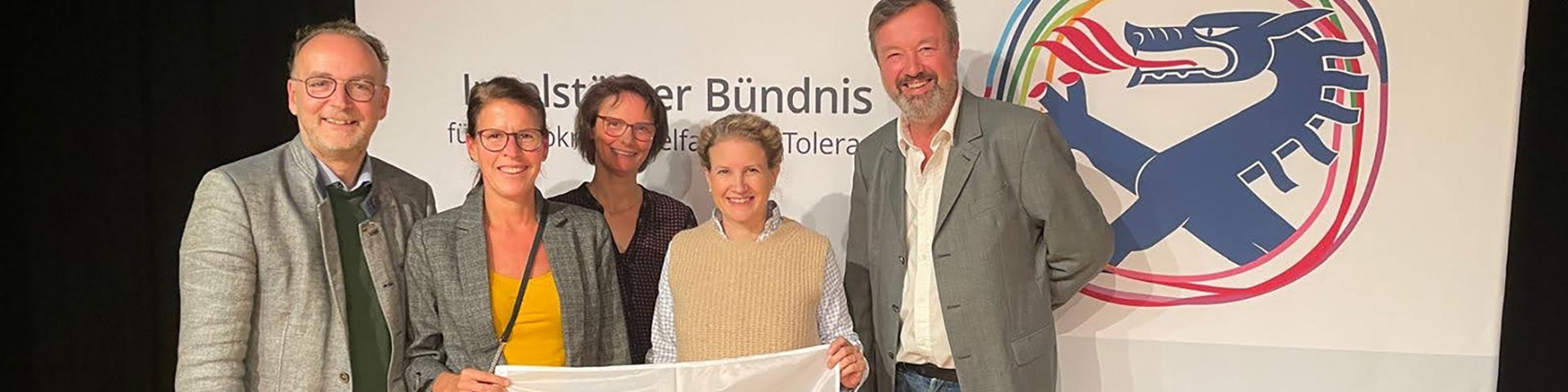 Fünf Personen stehen vor dem Logo des Ingolstädter Bündnisses und halten eine Fahne mit der Aufschrift ‚Zusammen Demokratie stärken!‘
