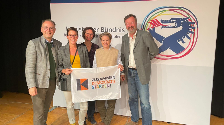 Fünf Personen stehen vor dem Logo des Ingolstädter Bündnisses und halten eine Fahne mit der Aufschrift ‚Zusammen Demokratie stärken!‘